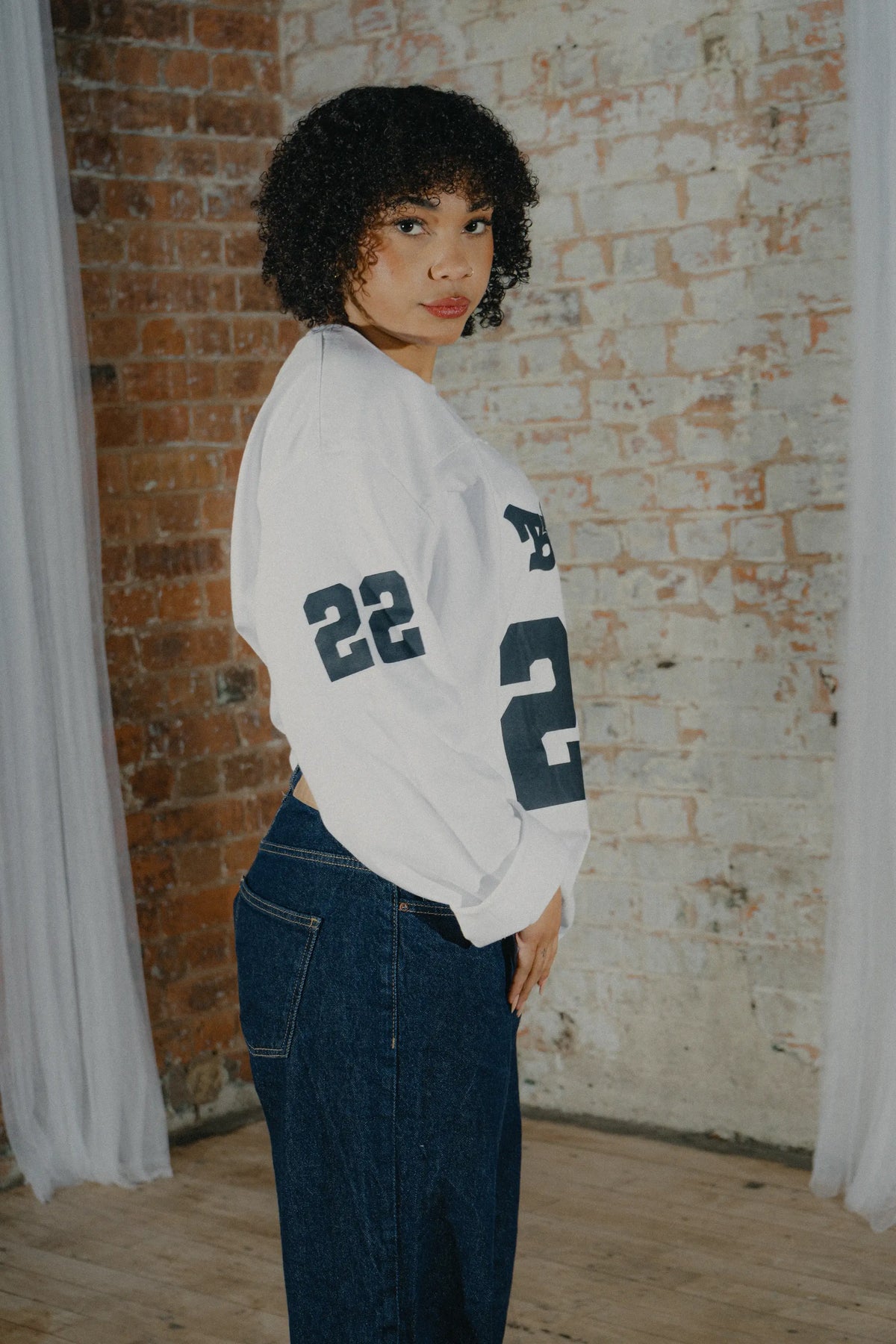 Woman modelling the BASICSS White 22 Long Sleeve Jersey with navy graphics, oversized fit and sleeve print, styled with dark blue denim jeans.
