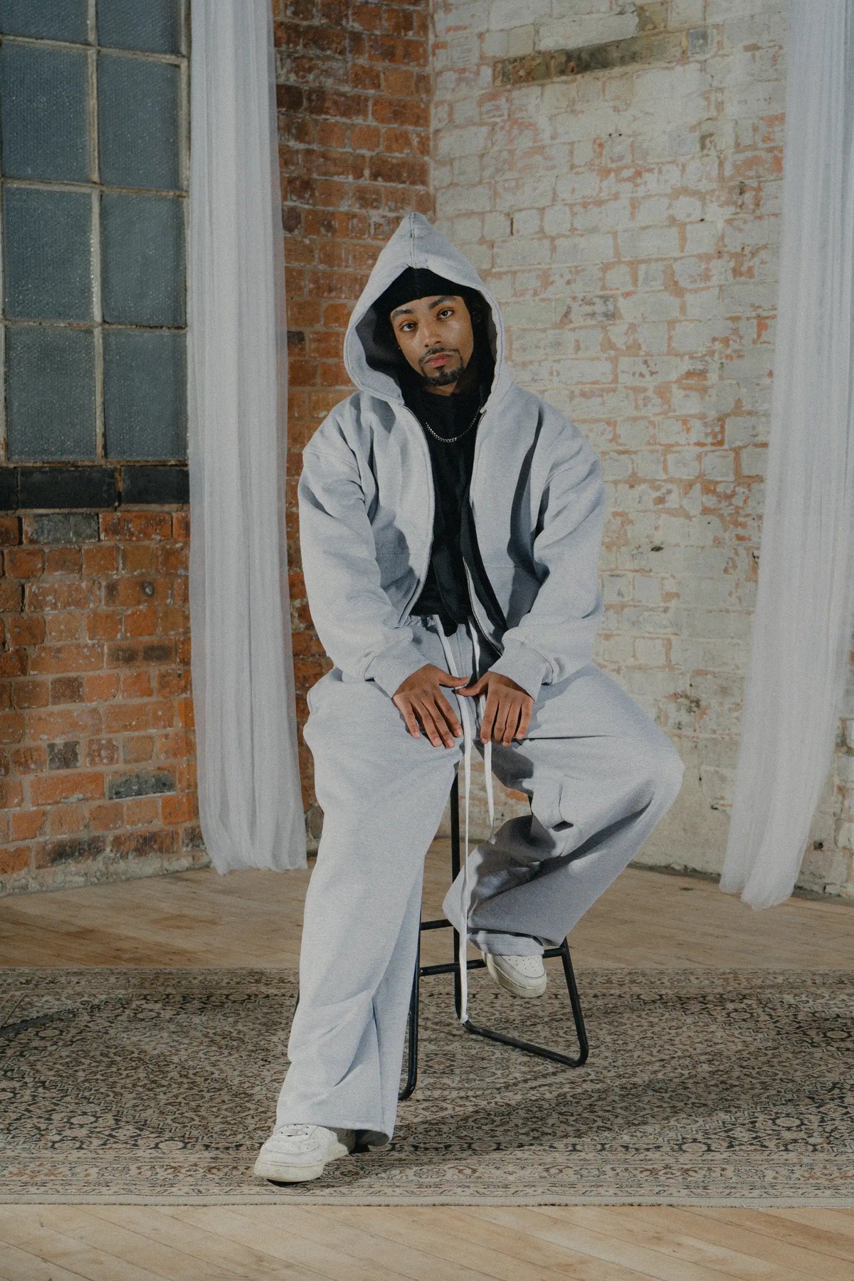 Man modelling the BASICSS Grey Cloud Tracksuit, featuring an oversized zip-up hoodie and matching heavyweight open-leg joggers, styled with white trainers in an industrial brick studio.