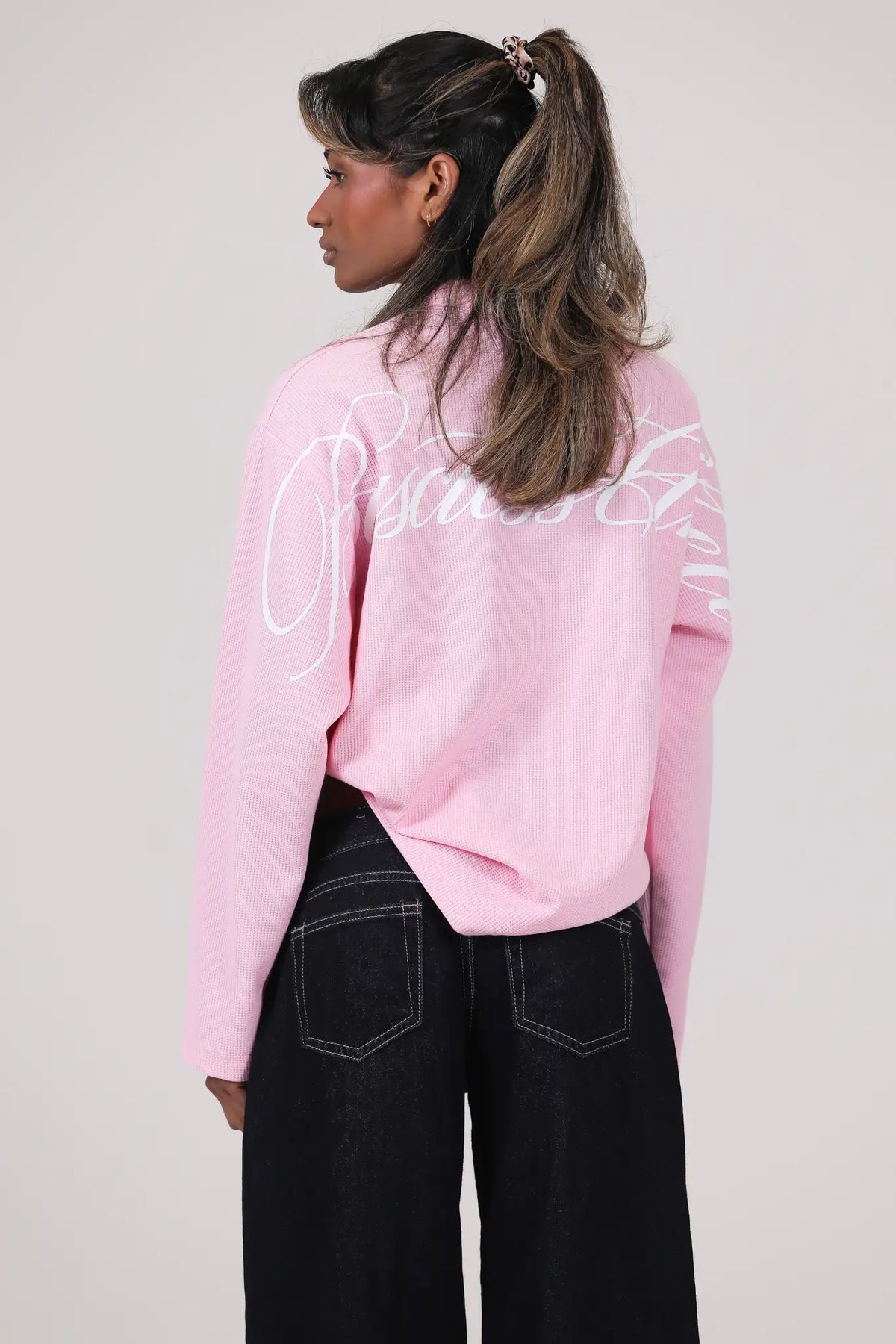 Woman modelling the BASICSS Pink Script Long Sleeve Jersey, featuring an oversized fit, textured fabric and large white cursive back print, styled with black wide-leg jeans.