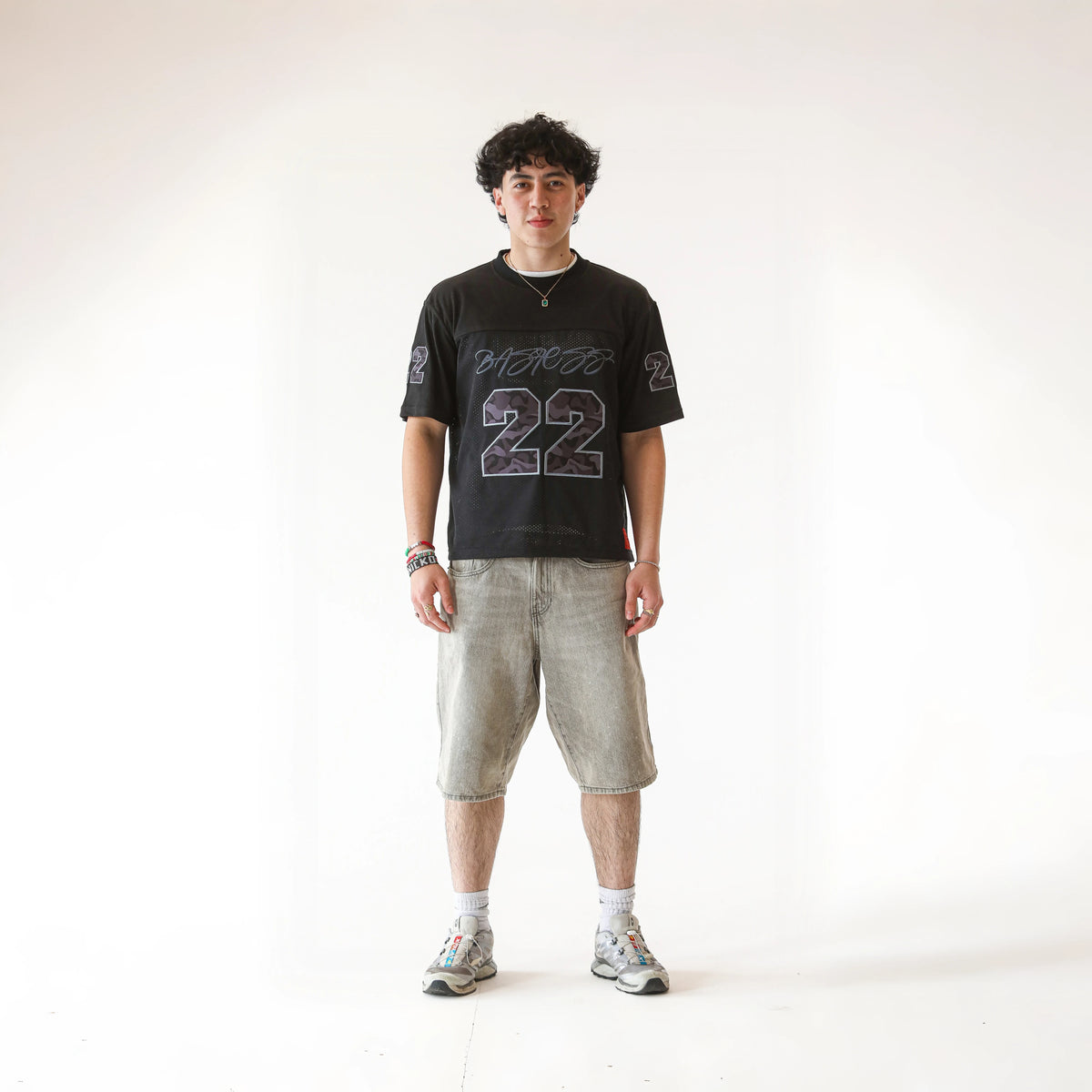 Man modelling the BASICSS Black 22 Camo Jersey, featuring an oversized fit, camouflage number 22 print and short sleeves, styled with grey denim shorts and trainers against a white studio background.
