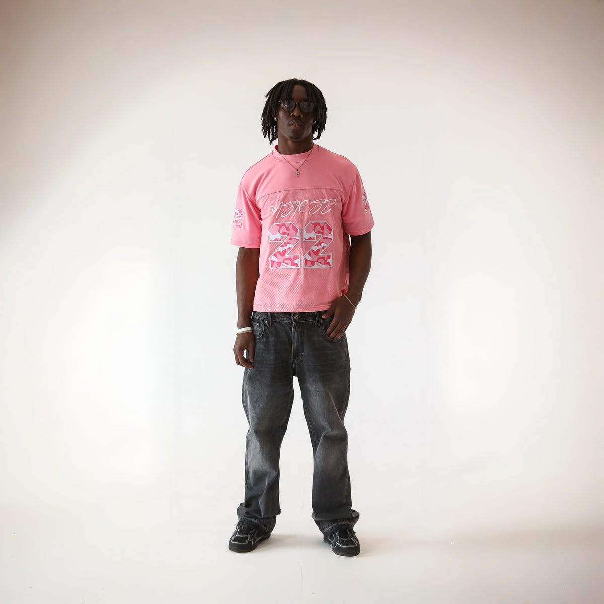 Man modelling the BASICSS Pink 22 Camo Jersey, featuring camouflage number graphics and short sleeves, styled with dark grey jeans, sunglasses and black shoes against a white studio background.
