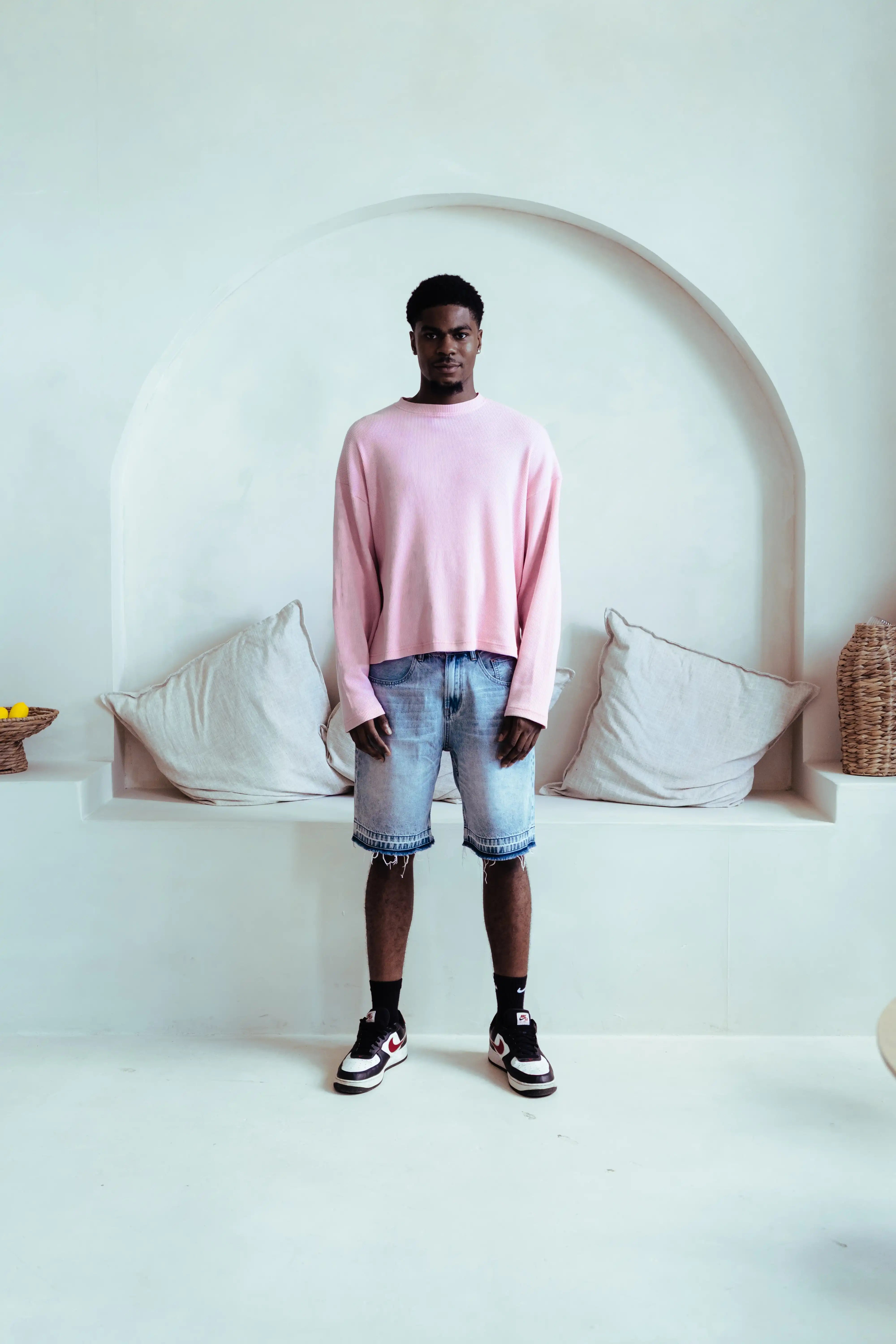 A man stands in a bright, minimalist white room with an arched alcove behind him, wearing a light pink long-sleeve knit sweater, light-wash denim shorts, black socks, and black-and-white sneakers.