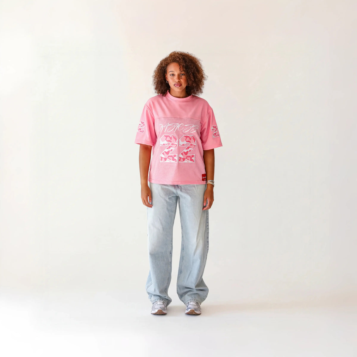Woman modelling the BASICSS Pink 22 Camo Jersey, featuring an oversized fit and camouflage number graphics, styled with light blue jeans and trainers against a white studio background.