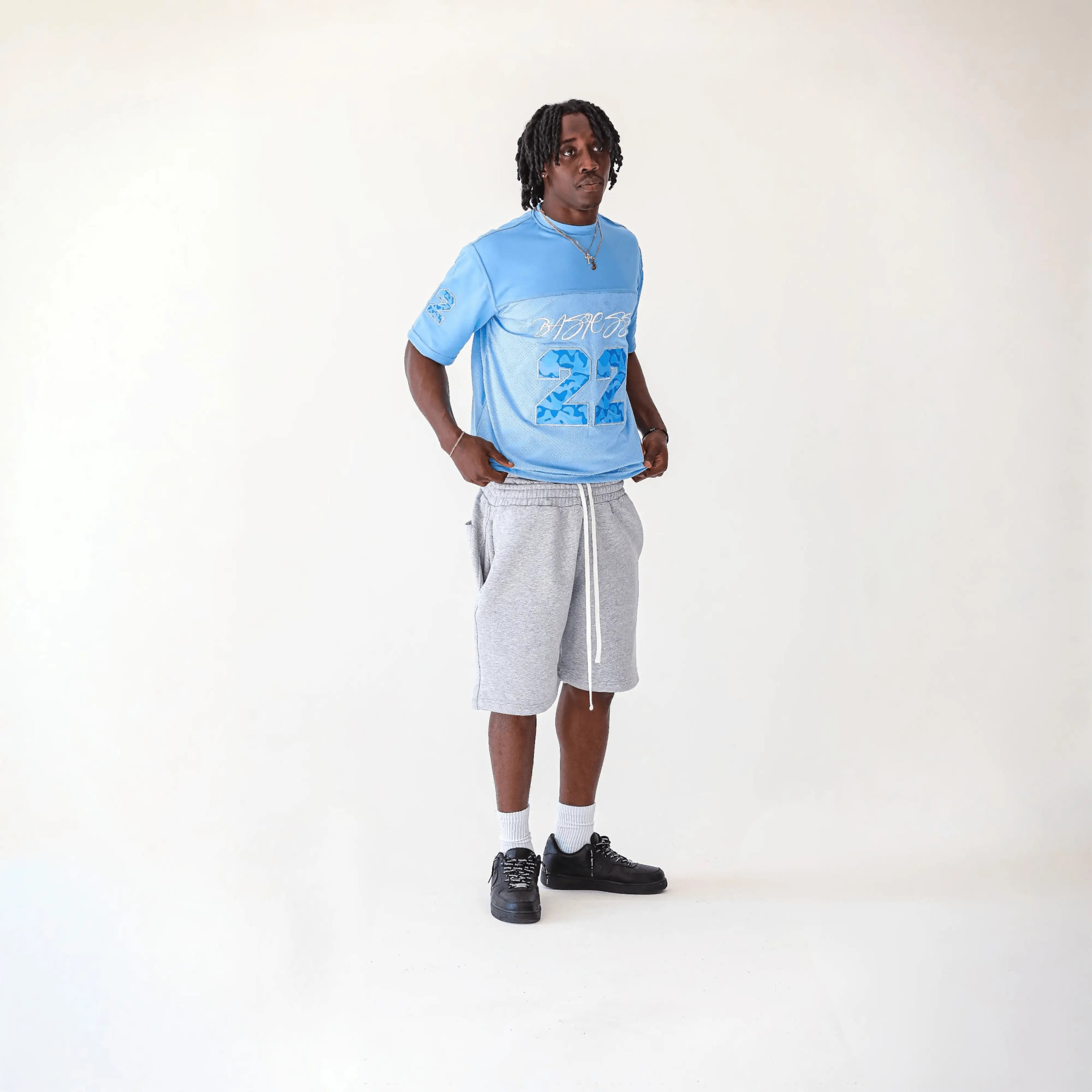 A man modeling a light blue graphic t-shirt, light gray sweat shorts, white socks, and black shoes against a plain off-white studio background.
