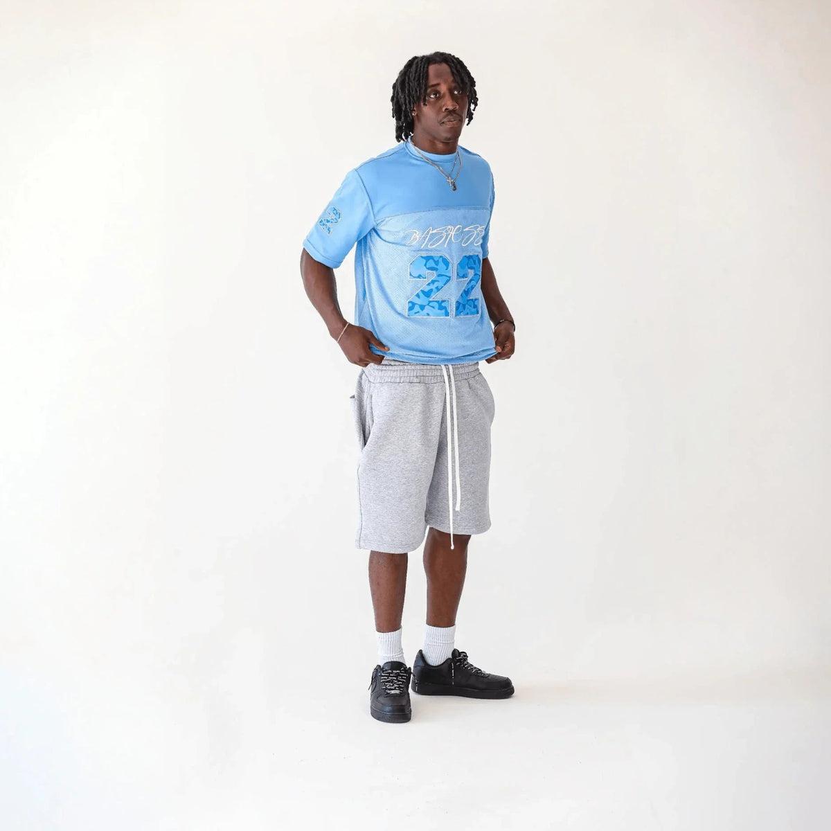 Man modelling the BASICSS Light Blue 22 Camo Jersey, featuring blue camouflage numbers and short sleeves, styled with grey shorts and black trainers against a white studio background.