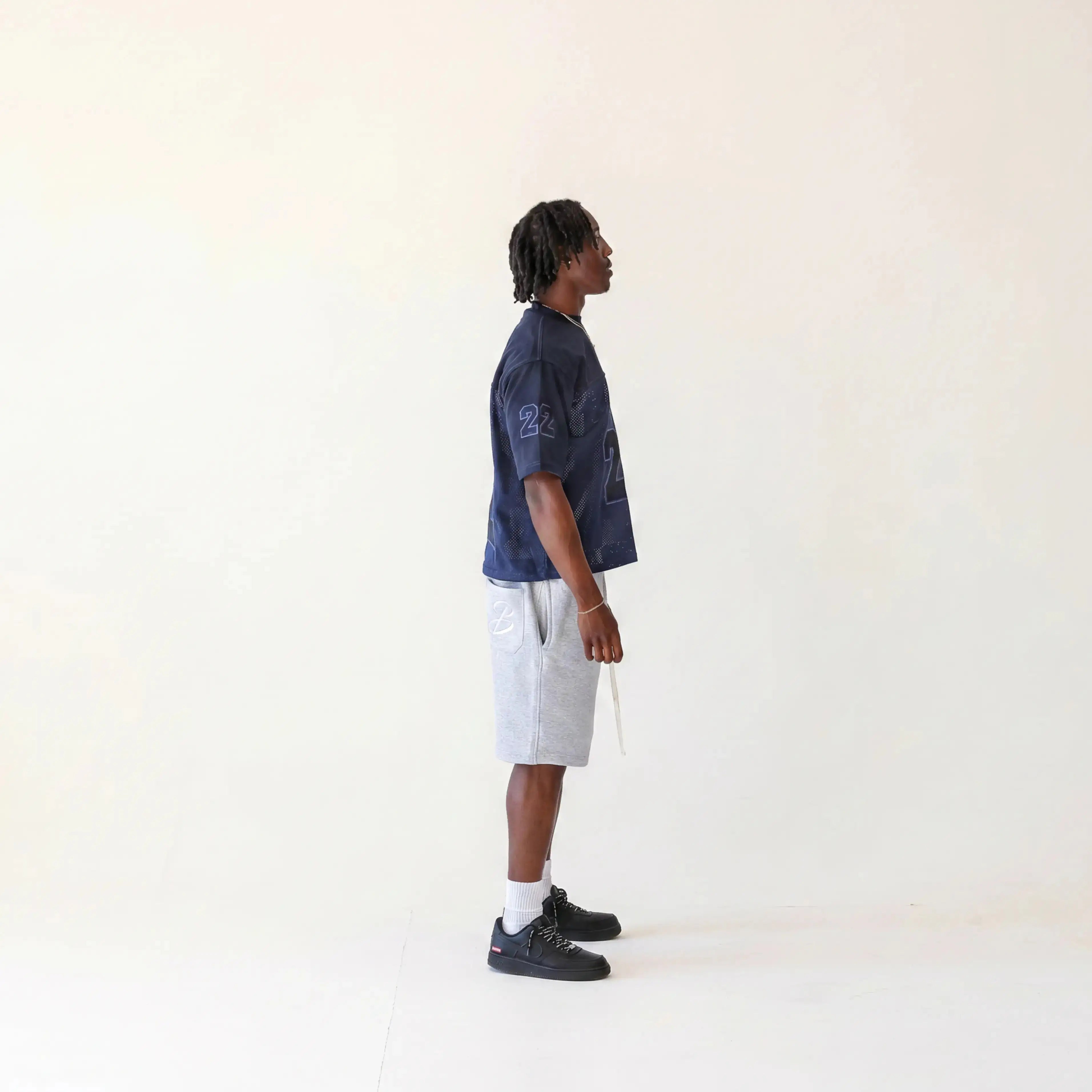 A man stands in profile wearing a dark navy short-sleeve jersey light grey shorts, white socks, and black sneakers against a plain off-white studio background.