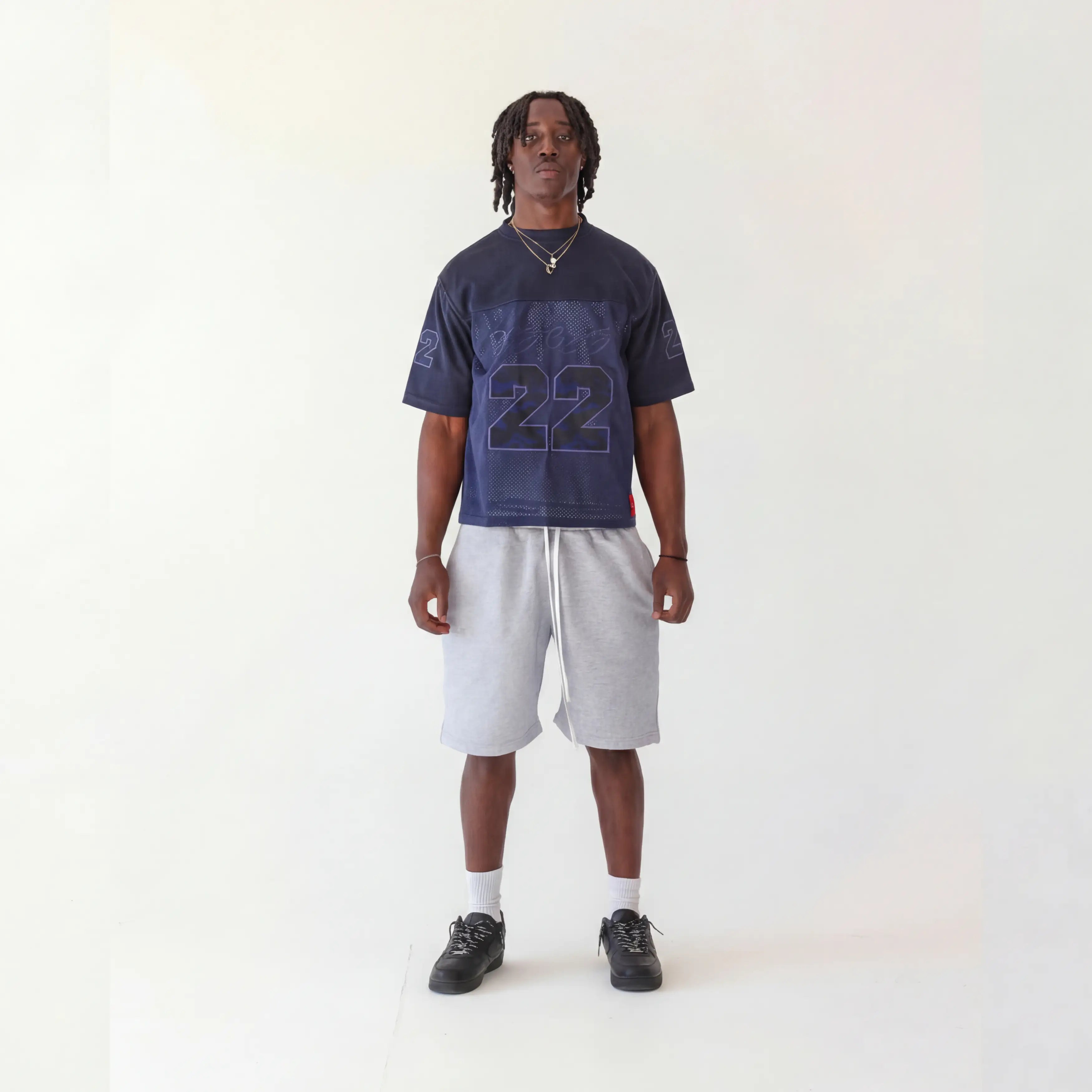 A man stands against a plain off-white studio background wearing a navy blue graphic t-shirt with a large number 22 on the front, light gray shorts, white socks, and black sneakers.