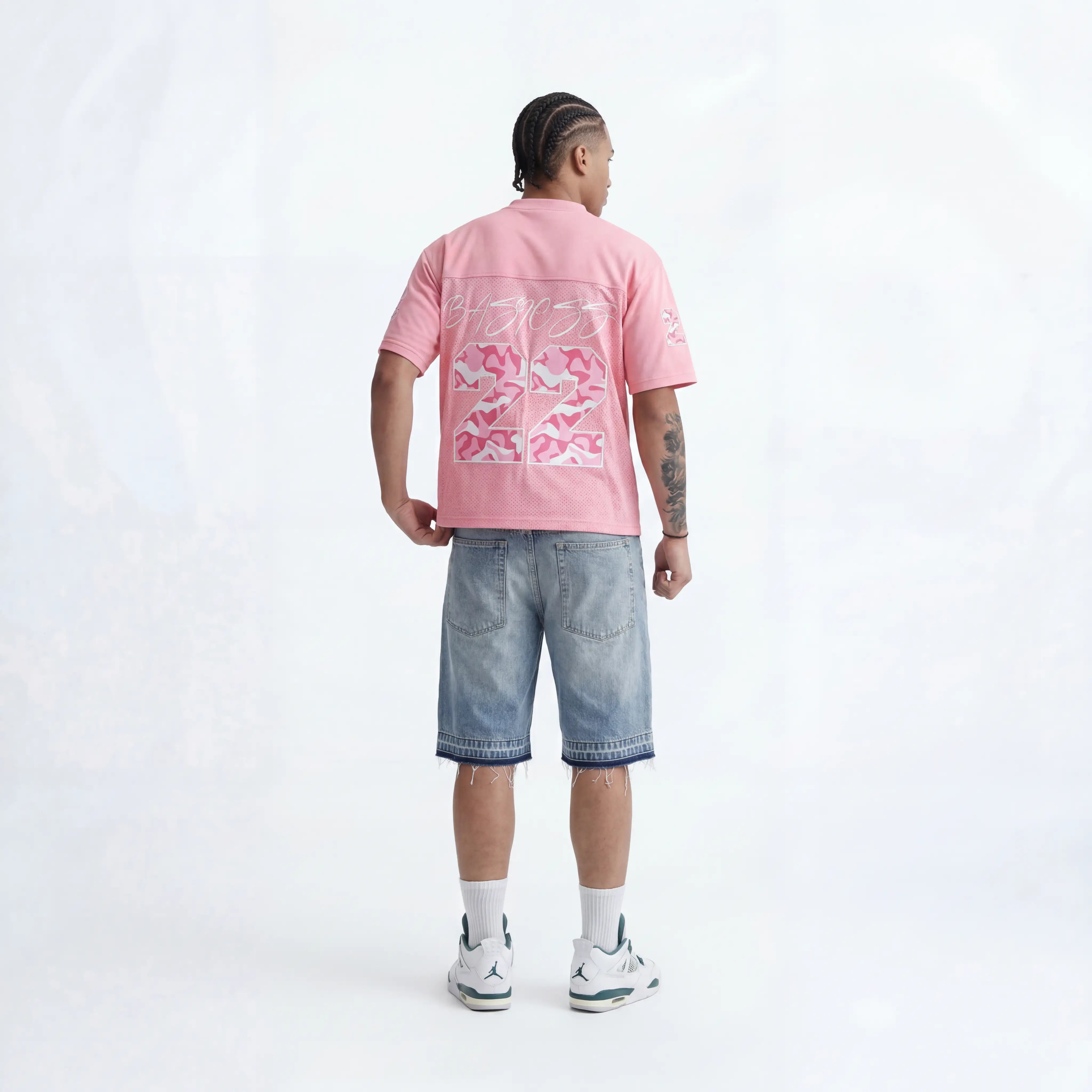 A man shown from the back wearing a pink t-shirt with a large pink and white graphic print, paired with light-wash denim shorts, white socks, and white sneakers with green accents, against a light gray studio background.