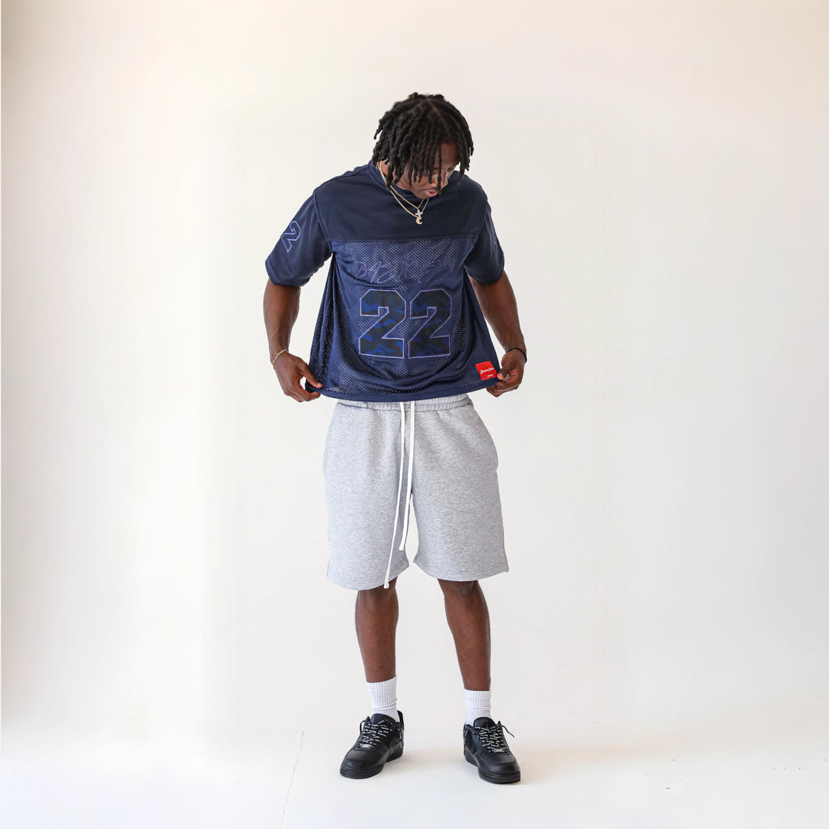 Man modelling the BASICSS Navy 22 Camo Jersey, featuring an oversized fit and camouflage 22 graphic, styled with grey shorts and black trainers against a white studio background.