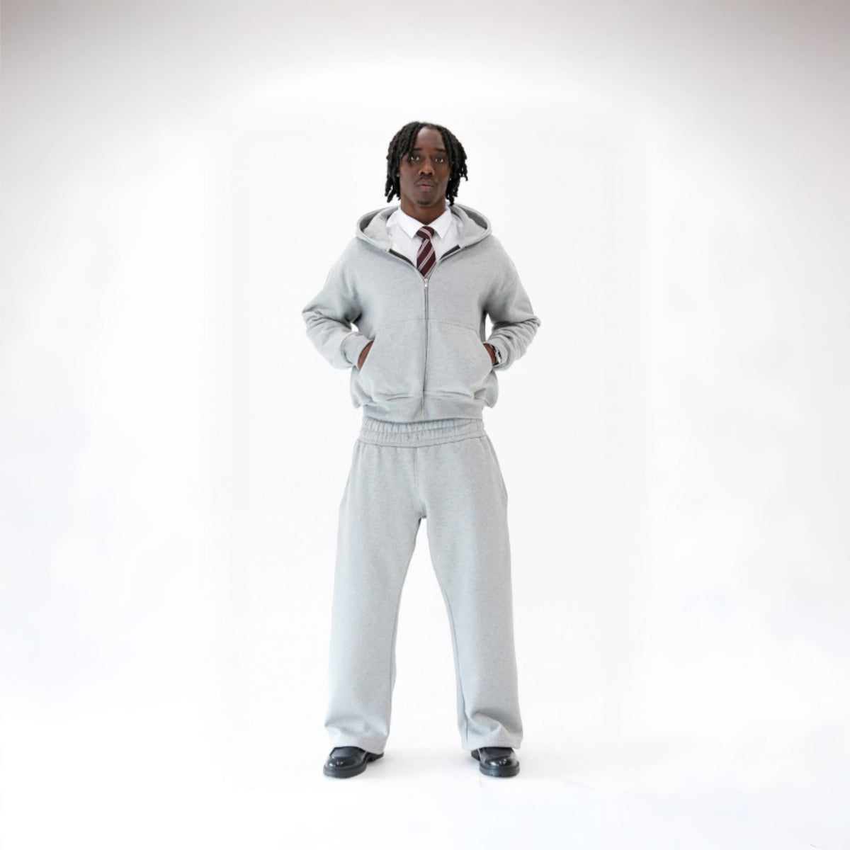 Man modelling the BASICSS Grey Cloud Tracksuit, featuring an oversized zip-up hoodie and matching heavyweight open-leg joggers, styled with a shirt, tie and black loafers against a white studio background.