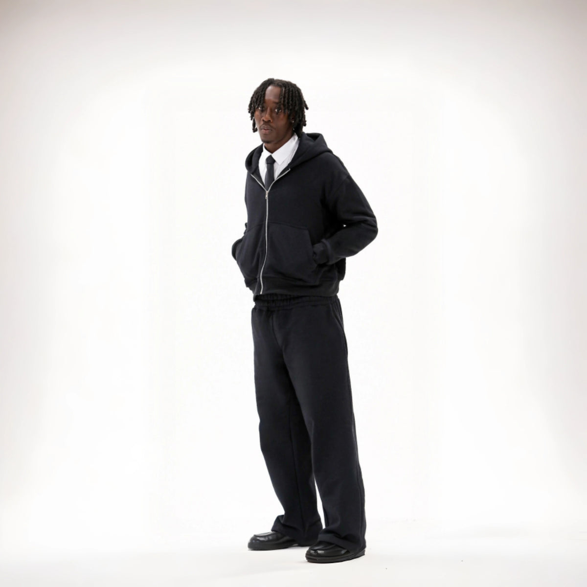 Man modelling the BASICSS Black Cloud Tracksuit, featuring an oversized zip-up hoodie, matching heavyweight open-leg joggers and deep front pockets, styled with a shirt, tie and black loafers against a white studio background.