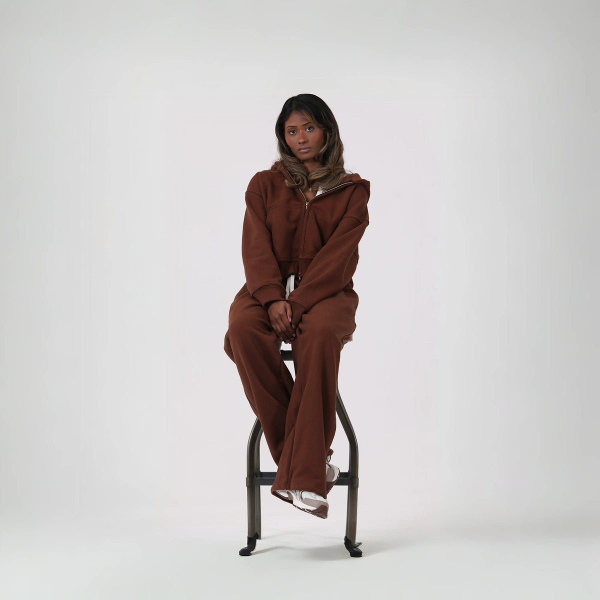 Woman seated on a stool wearing the BASICSS Mocha Cloud Tracksuit, featuring a zip-up hoodie and matching open-leg joggers, styled with trainers against a light studio background.