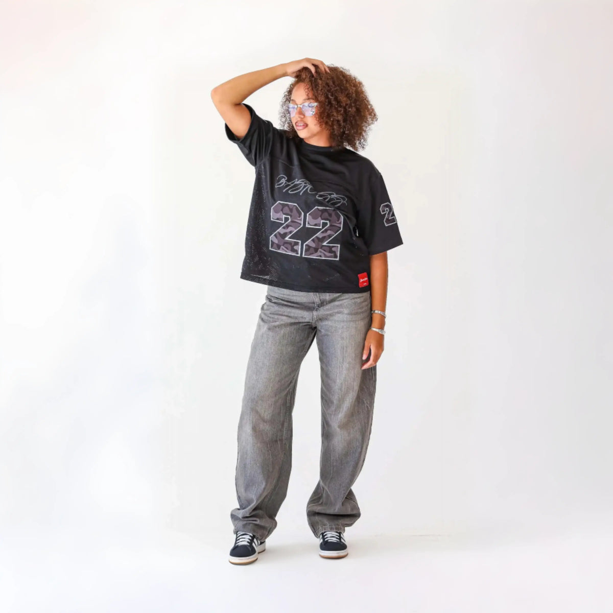 Woman modelling the BASICSS Black 22 Camo Jersey, featuring an oversized fit and camouflage number graphics, styled with grey jeans, trainers and sunglasses against a white studio background.