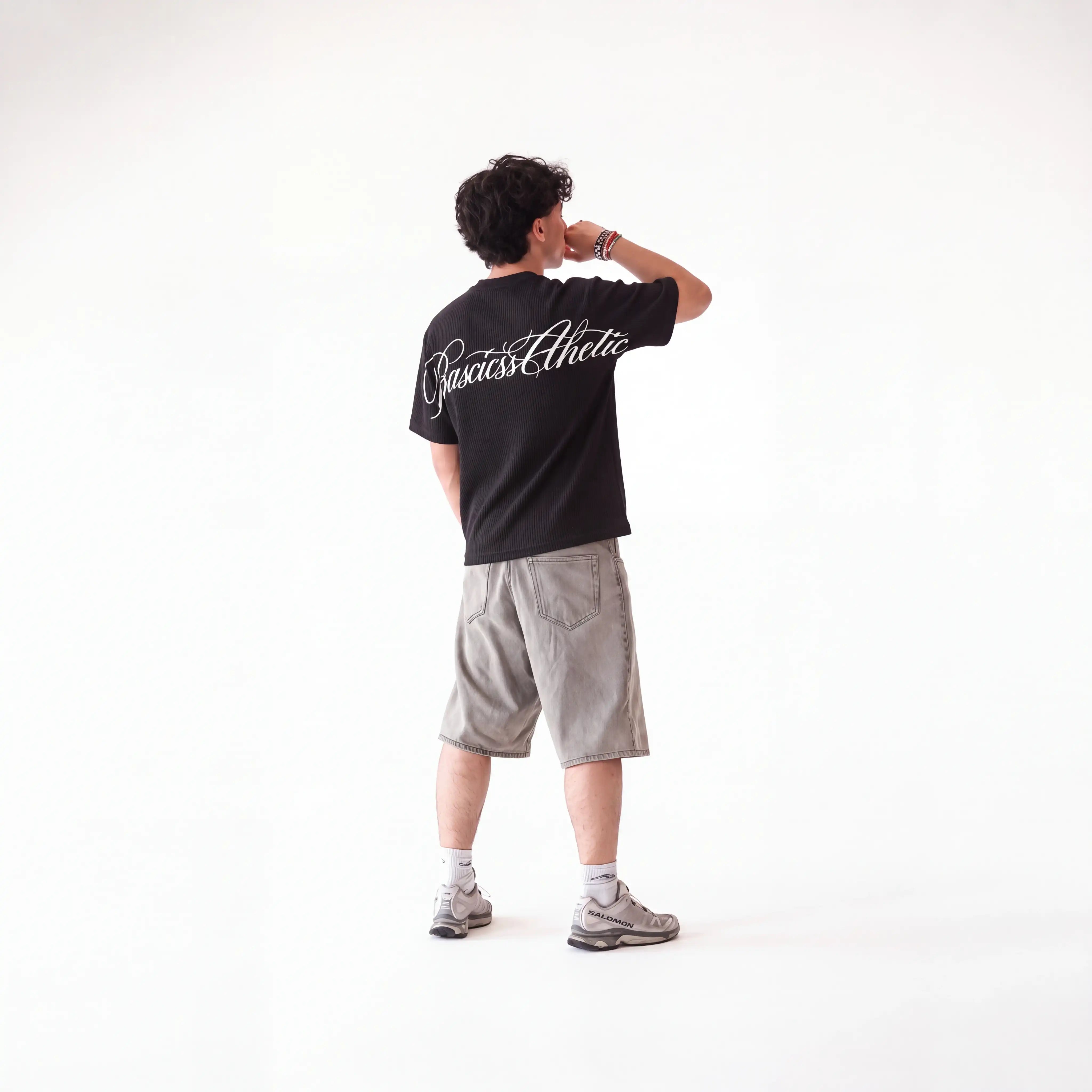 A man standing with his back to the camera wearing a black t-shirt with 'Basicss Athletic' printed in white script across the upper back, paired with gray shorts, white socks, and gray athletic shoes, against a plain white studio background.