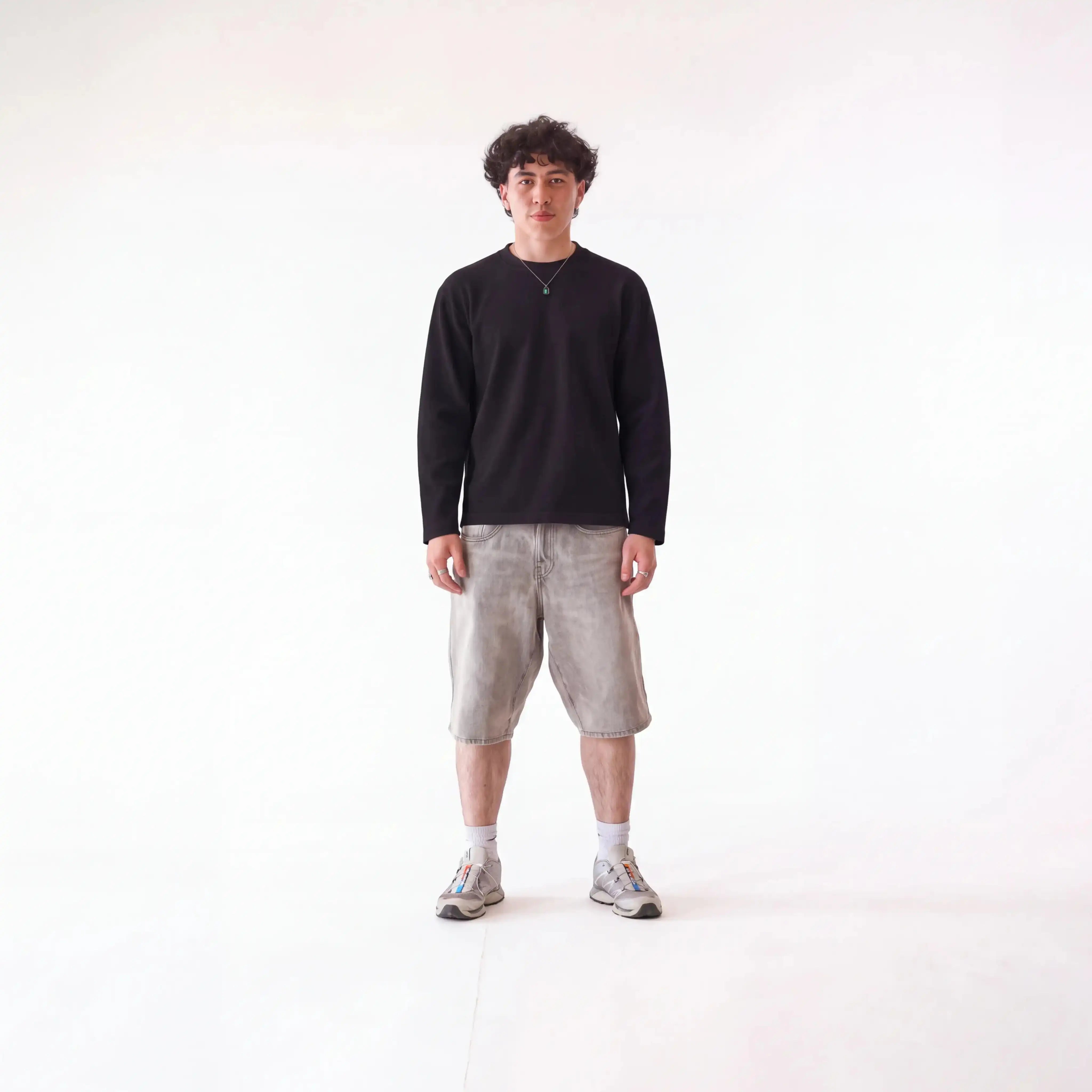 A man stands against a seamless white studio background wearing a black long-sleeve crewneck shirt, light beige corduroy shorts, white socks, and gray sneakers.