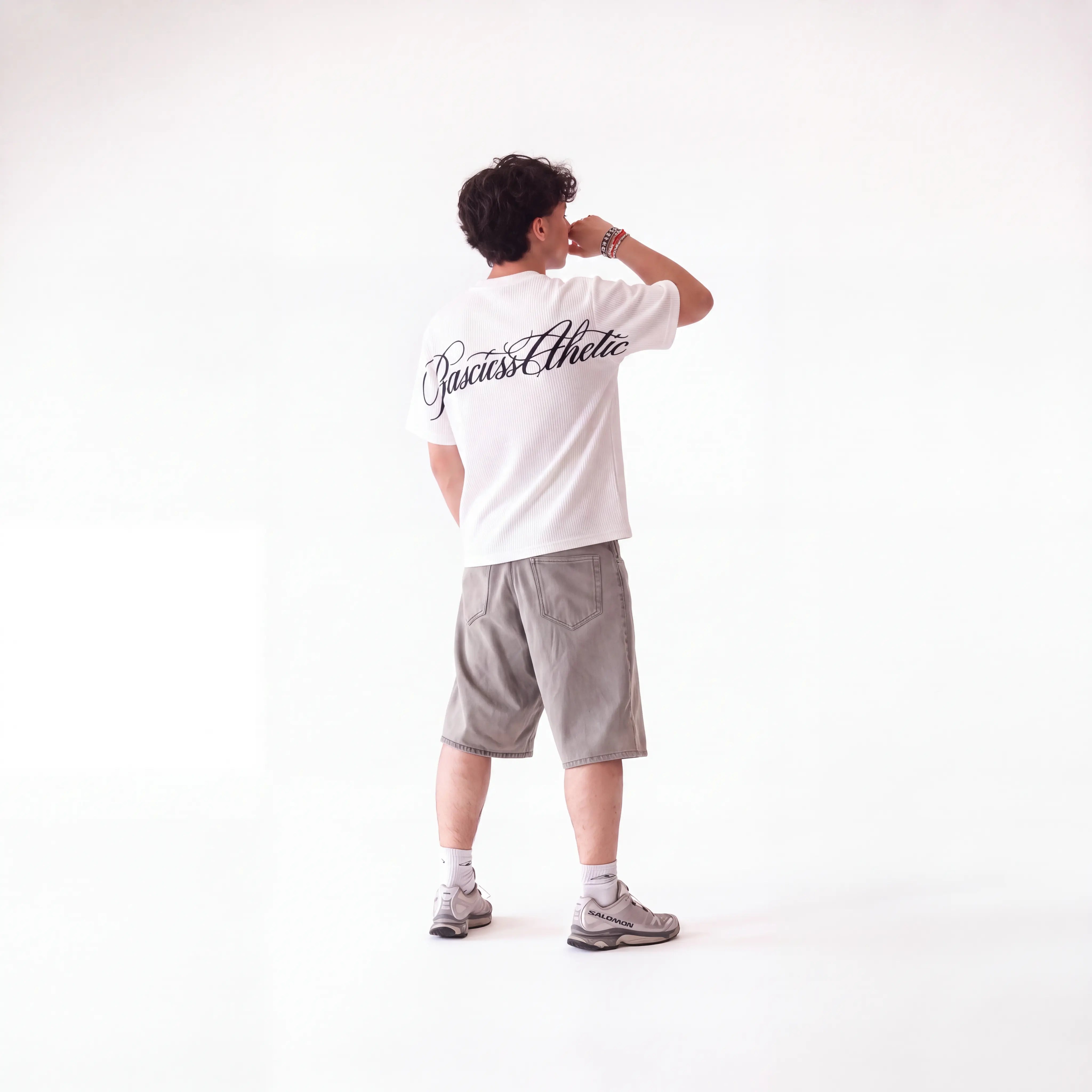 A man standing with his back to the camera wearing a white t-shirt with 'Basicss Athletic' printed in black script on the back, paired with gray shorts and gray sneakers, against a plain white studio background.