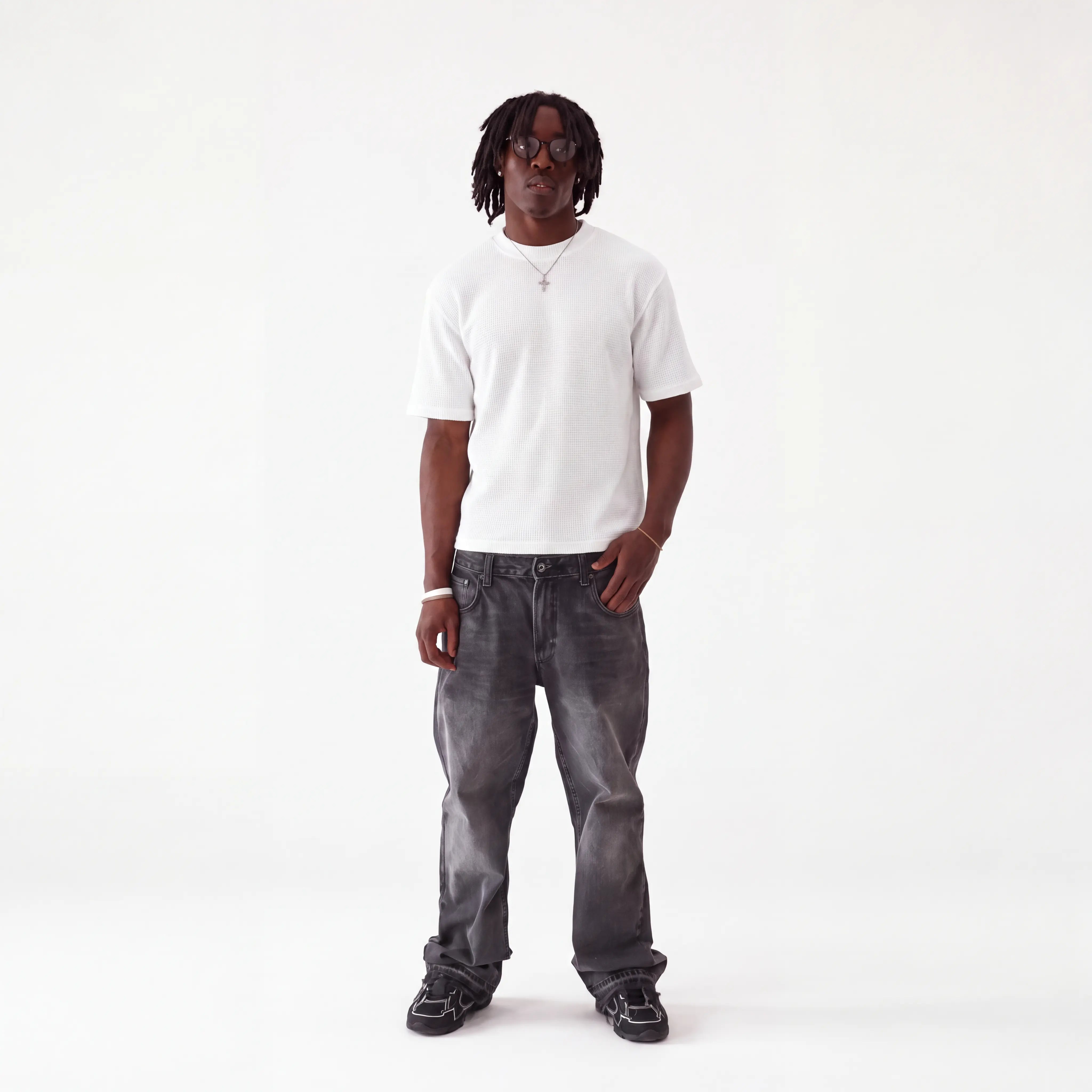 A man wearing a white crew-neck t-shirt, dark gray washed jeans, and black sneakers, standing against a plain white studio background.