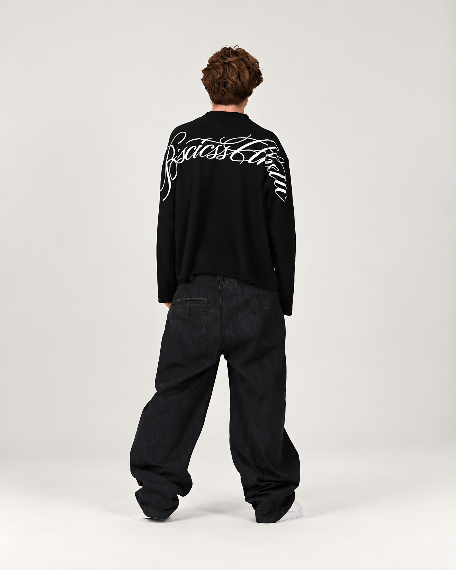 A model viewed from the back wearing a black long-sleeve shirt with a large white script logo across the upper back, paired with loose black jeans, against a plain light gray studio background.
